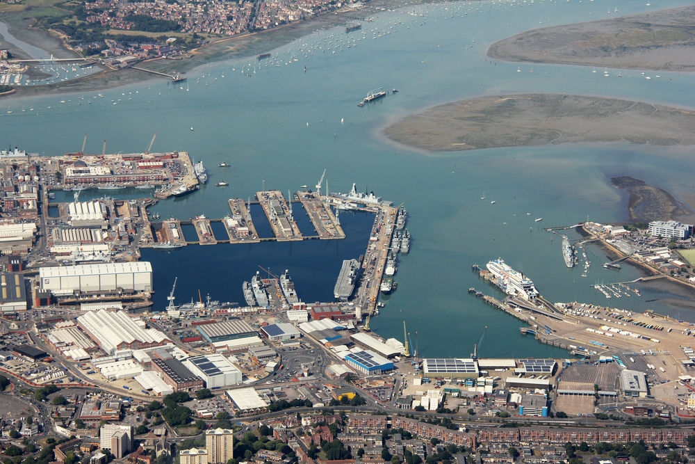 Portsmouth Harbour Naval Base, with an aircraft carrier in the inner harbour and various other grey-painted warships tied up.
